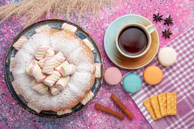 Top view of sugar powdered cake with macarons and cup of tea on pink surface