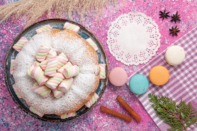 Top view of sugar powdered cake with macarons and marshmallow on pink surface