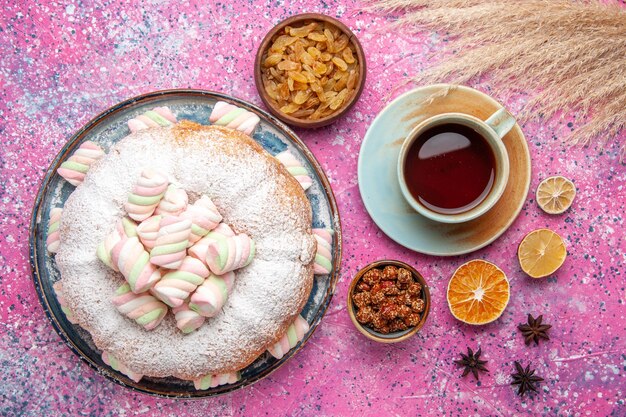 Top view of sugar powdered cake with sweet marshmallows and cup of tea on pink surface