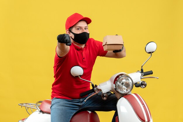 Вид сверху на доставщика Sumiling в красной блузке и шляпных перчатках в медицинской маске, сидящего на скутере, показывая порядок, указывающий вперед