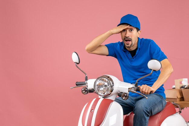 Top view of surprised courier guy wearing hat sitting on scooter delivering orders on pastel peach