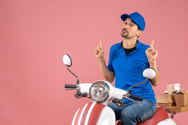 Top view of surprised courier guy wearing hat sitting on scooter delivering orders pointing up on pastel peach