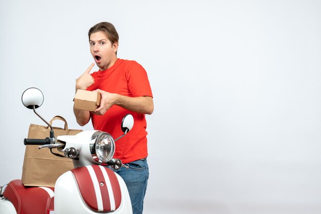 Top view of surprised delivery guy in red uniform standing near scooter on white wall