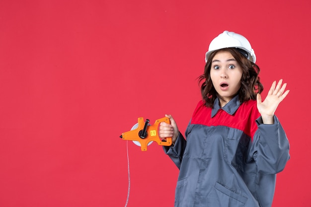 Top view of surprised emotional female architect in uniform with hard hat opening measuring tape on isolated red background