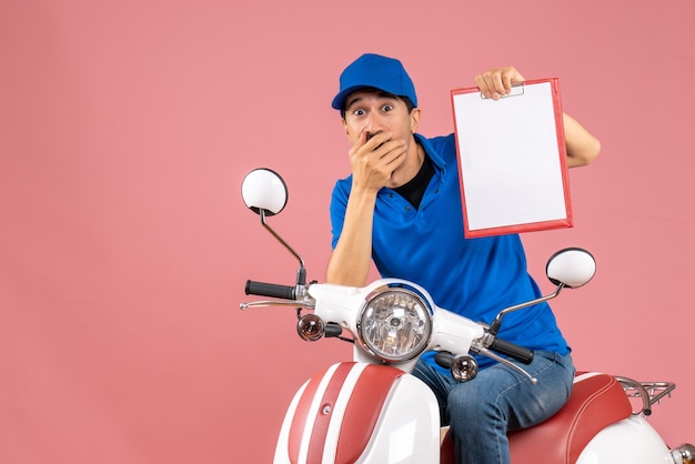 Top view of surprised shocked courier man wearing hat sitting on scooter holding document on pastel peach