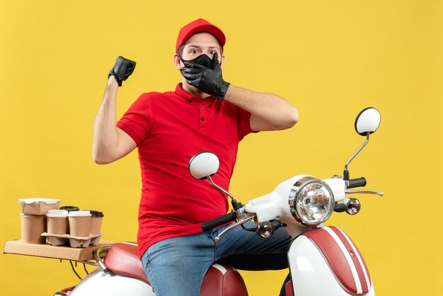 Top view of surprised young adult wearing red blouse and hat gloves in medical mask delivering order sitting on scooter pointing back on yellow wall