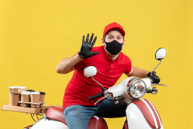 Top view of surprised young adult wearing red blouse and hat gloves in medical mask delivering order sitting on scooter showing five on yellow wall