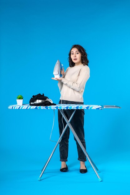 Top view of surprised young girl standing behind the board and holding iron on blue wave background