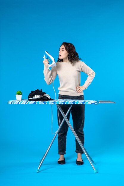 Top view of surprised young girl standing behind the board and holding iron checking it on blue wave background