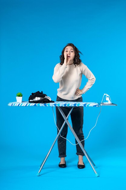 Top view of surprised young girl standing behind the iron board and calling someone on blue wave background