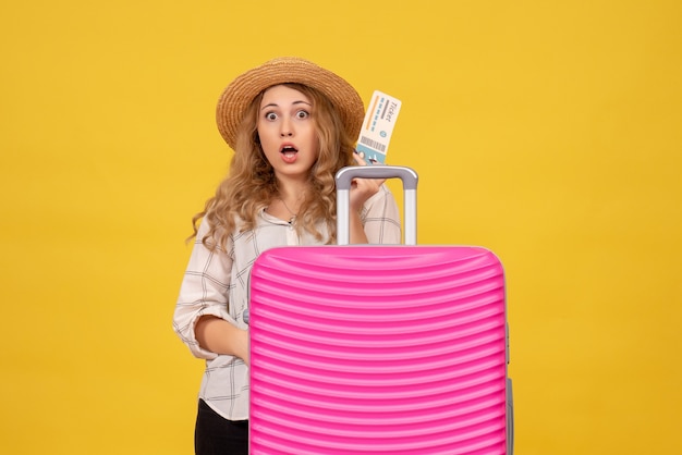 Top view of surprised young lady wearing hat holding ticket and standing behind her pink bag