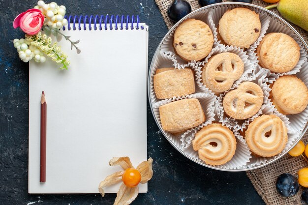 ダークブルーのデスクにさまざまな新鮮なフルーツと空白のメモ帳と一緒にトップビューの甘いクッキーフルーツクッキービスケット甘い新鮮な