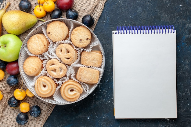 ダークブルーのデスクにさまざまな新鮮なフルーツのメモ帳と一緒にトップビューの甘いクッキーフルーツクッキービスケット甘い新鮮な