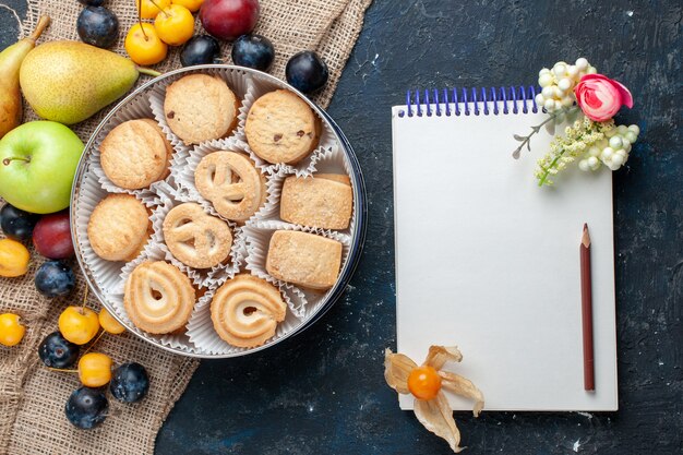 Top view sweet cookies along with different fresh fruits notepad on the dark-blue desk fruit cookie biscuit sweet fresh