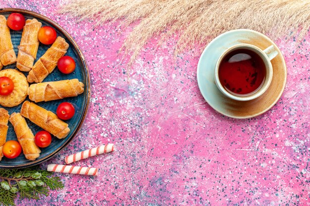 Вид сверху сладкие вкусные рогалики внутри подноса со свежими кислыми сливами и чаем на светло-розовом столе