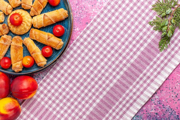 Top view sweet delicious bagels inside tray with plums and peaches on light-pink desk