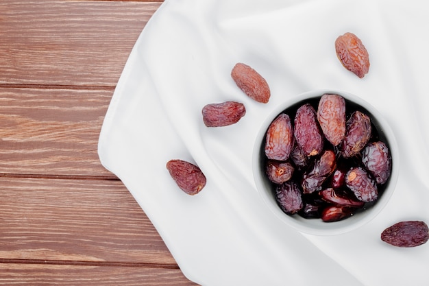 Top view of sweet dried date fruits in a bowl on white tablecloth on wooden background with copy space