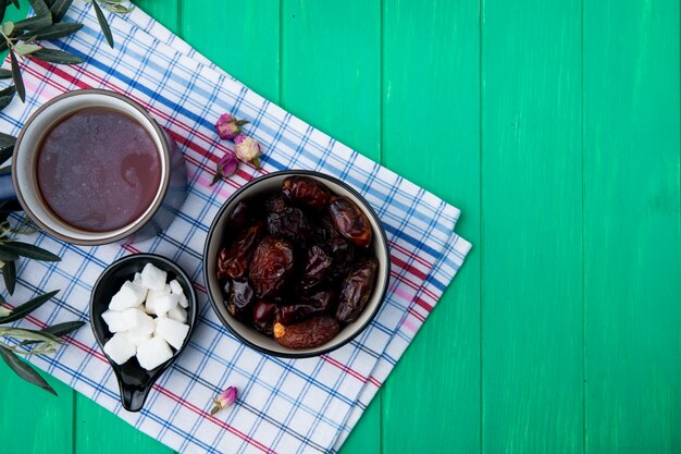 Вид сверху сладких сушеных фиников в миске с чашкой чая и кусочками сахара в блюдце на плед скатерть на зеленой древесине с копией пространства