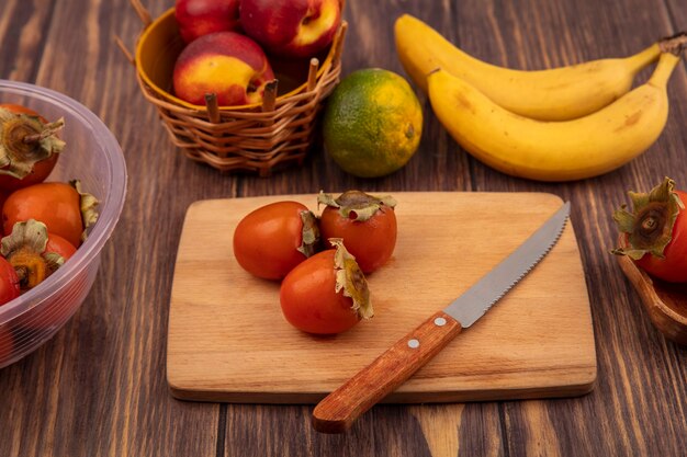 Top view of sweet persimmons on a wooden kitchen board with knife with peaches on a bucket with tangerine and bananas isolated on a wooden background