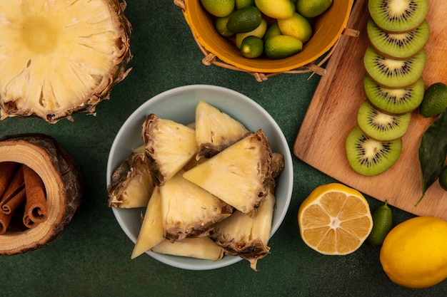 緑の背景に分離されたレモンとシナモンスティックとバケツにキンカンと木製のキッチンボードにキウイスライスとボウルに甘いパイナップルスライスの上面図
