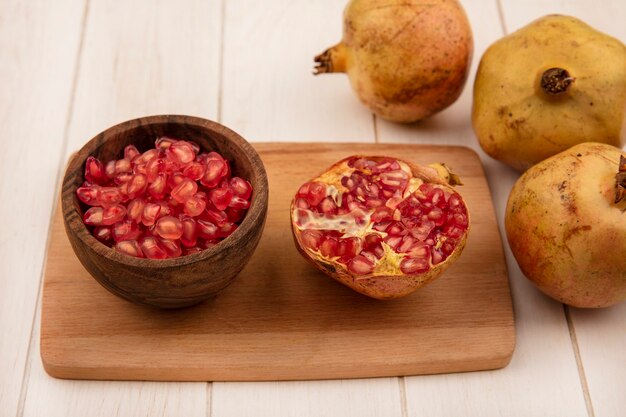 Top view of sweet pomegranate seeds on a wooden bowl on a wooden kitchen board with pomegranates isolated on a white wooden wall