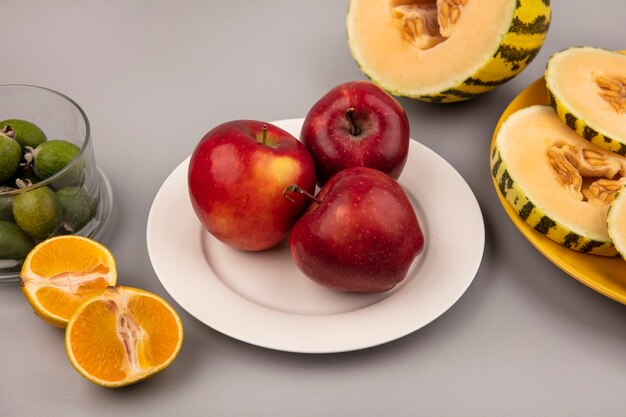 Top view of sweet red apples on a white plate with slices of cantaloupe melon on a yellow plate with tangerines isolated on a grey wall