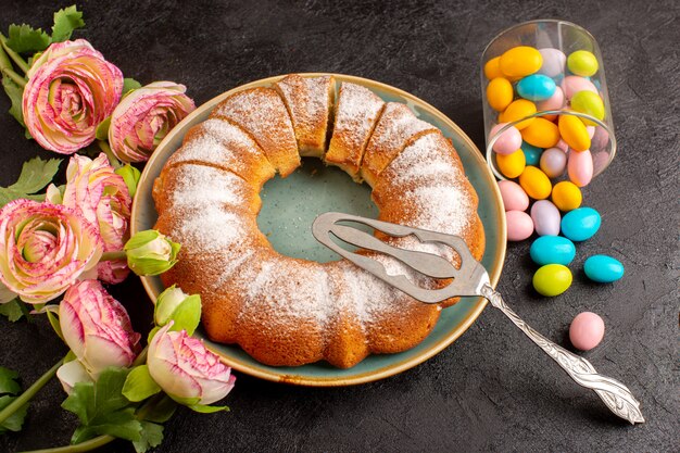 A top view sweet round cake with sugar powder along with colorful candies sliced sweet delicious isolated cake inside plate and grey background biscuit sugar cookie