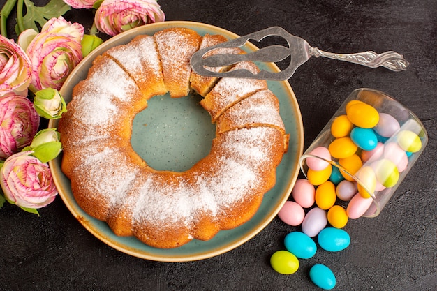 A top view sweet round cake with sugar powder along with colorful candies sliced sweet delicious isolated cake inside plate and grey background biscuit sugar cookie