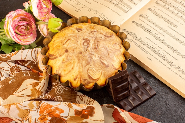 Вид сверху сладкий круглый торт вкусный вкусный внутри формы для кекса вместе с шоколадными батончиками цветы и тетрадь нот на сером фоне печенье сахарное печенье