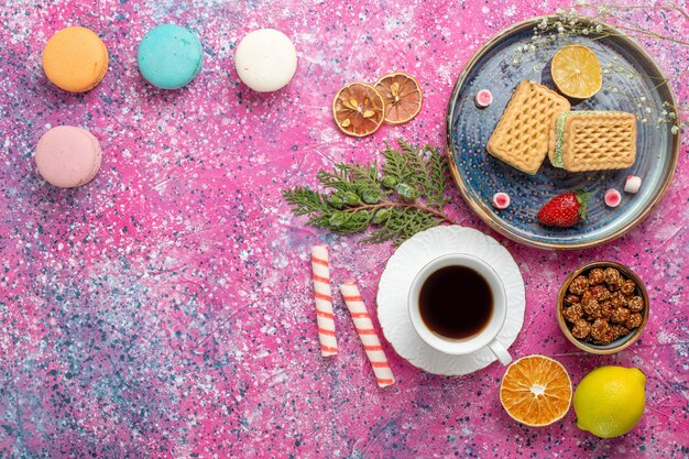 Top view of sweet waffles with cup of tea and macarons on the pink surface