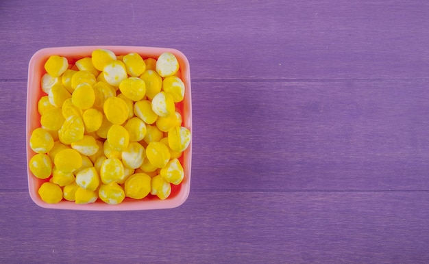 Top view of sweet yellow candies in a bowl on purple wooden background with copy space