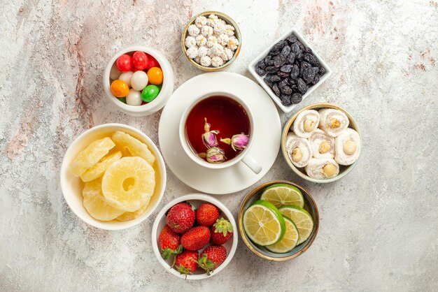 Top view sweets in bowls a cup of black tea on the saucer between bowls of straberries slices of limes and sweets on the white table