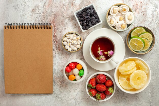 テーブルの右側にあるクリーム色のノートと紅茶のカップの横にあるライム乾燥パイナップルとお菓子のボウルのトップビューのお菓子