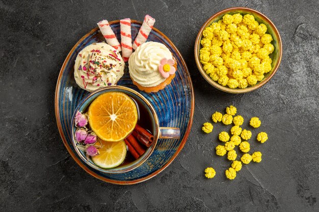 Вид сверху сладости на тарелке кексы с белым кремом на блюдце чашка чая с лимоном и палочками корицы миска желтых конфет на столе