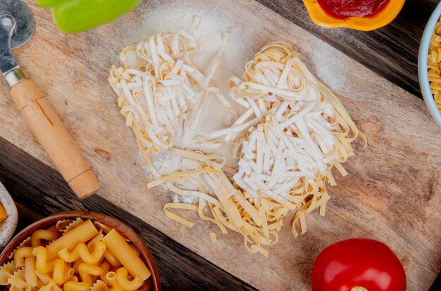 Top view of tagliatelle macaroni with flour pepper and tomato on cutting board with other types ketchup on wooden surface