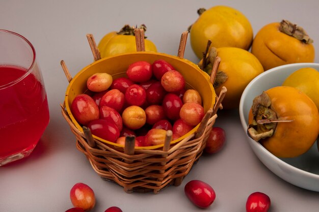 Top view of tart sweet cornelian fruits on a bucket with persimmon fruits on a bowl with fresh cornelian fruit juice in a glass on a grey surface