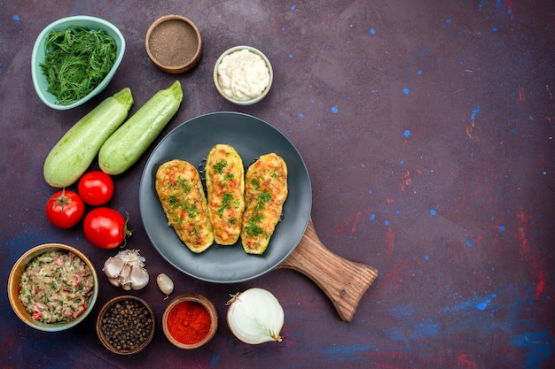 Top view of tasty baked squashes with greens along with fresh vegetables and seasonings on dark surface