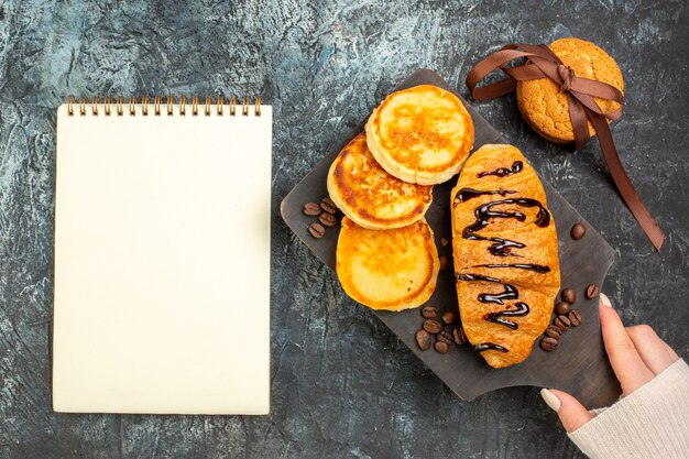 Top view of tasty breakfast with pancakes croisasant stacked cookies and notebook on dark surface