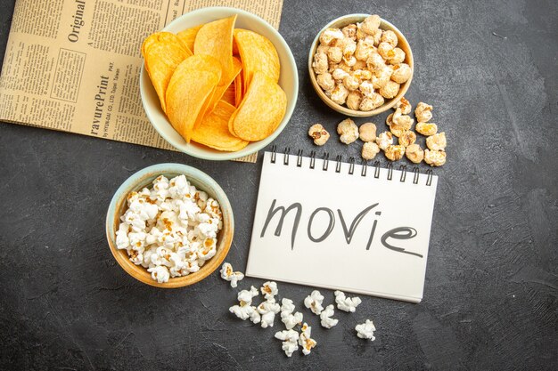 Top view tasty cheese chips with different snacks and movie written notepad on dark background