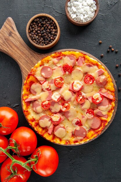Вид сверху вкусной сырной пиццы с сосисками и помидорами на темном столе