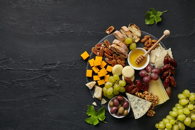 Top view of tasty cheese plate with fruit, grape, nuts and honey on black desk.