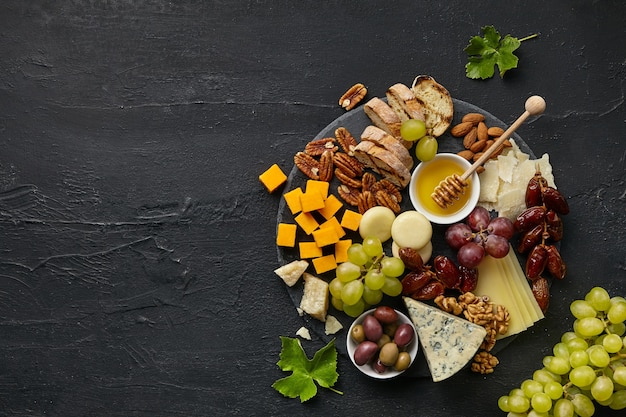 Top view of tasty cheese plate with fruit, grape, nuts and honey on black desk.