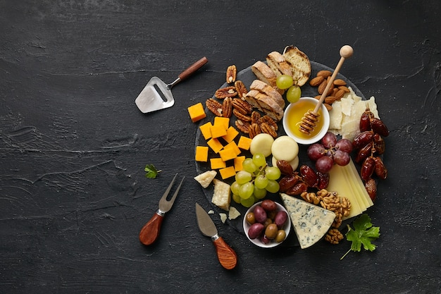Top view of tasty cheese plate with fruit, grape, nuts and honey on a circle kitchen plate on the black stone background, top view, copy space. Gourmet food and drink.