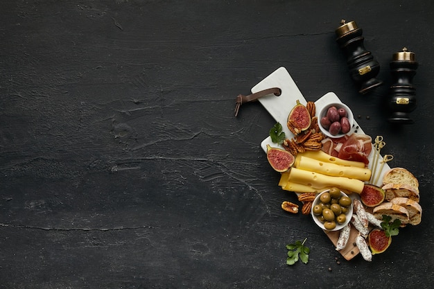 Top view of tasty cheese plate with fruit, grape, nuts, olives, bacon and toasted bread on a wooden kitchen plate on the black stone background, top view, copy space. Gourmet food and drink.