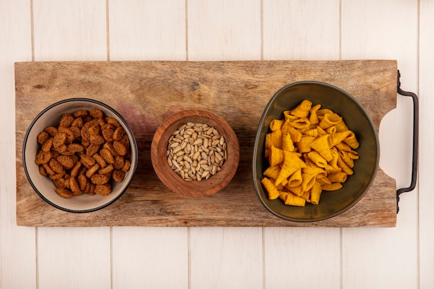 Top view of tasty cone shape fried corn snacks on a bowl with small rye rusks with shelled sunflower seeds on a wooden bowl on a wooden kitchen board on a beige wooden table