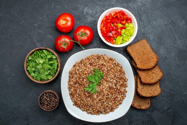 Top view tasty cooked buckwheat with dark bread loafs and greens on dark space