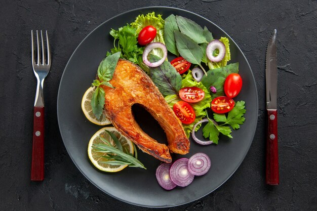 暗いテーブルの上に新鮮な野菜とカトラリーを添えたおいしい調理済み魚の上面図