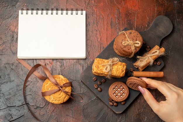 Top view tasty cookies tied with rope cinnamon sticks biscuits on wood serving board biscuit in female hand notepad on dark table