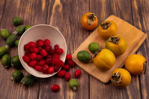 Free photo top view of tasty cornelian cherries falling out of a bowl with persimmon and feijoas on a wooden kitchen board on a wooden surface