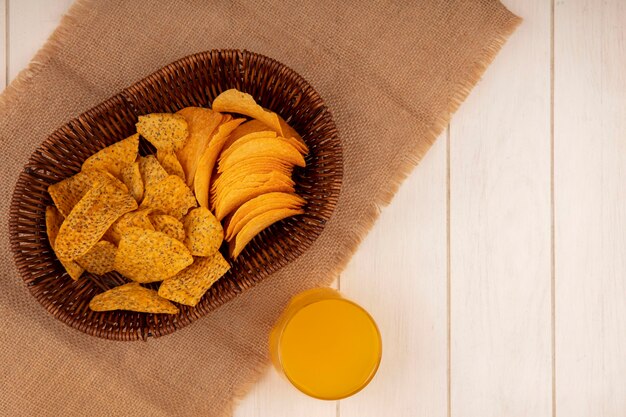 Top view of tasty crispy chips on a bucket on a sack cloth with a glass of orange juice on a beige wooden table with copy space